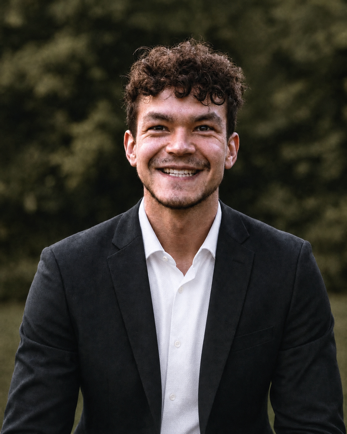 Luc smiling outdoors in a black jacket and white shirt
